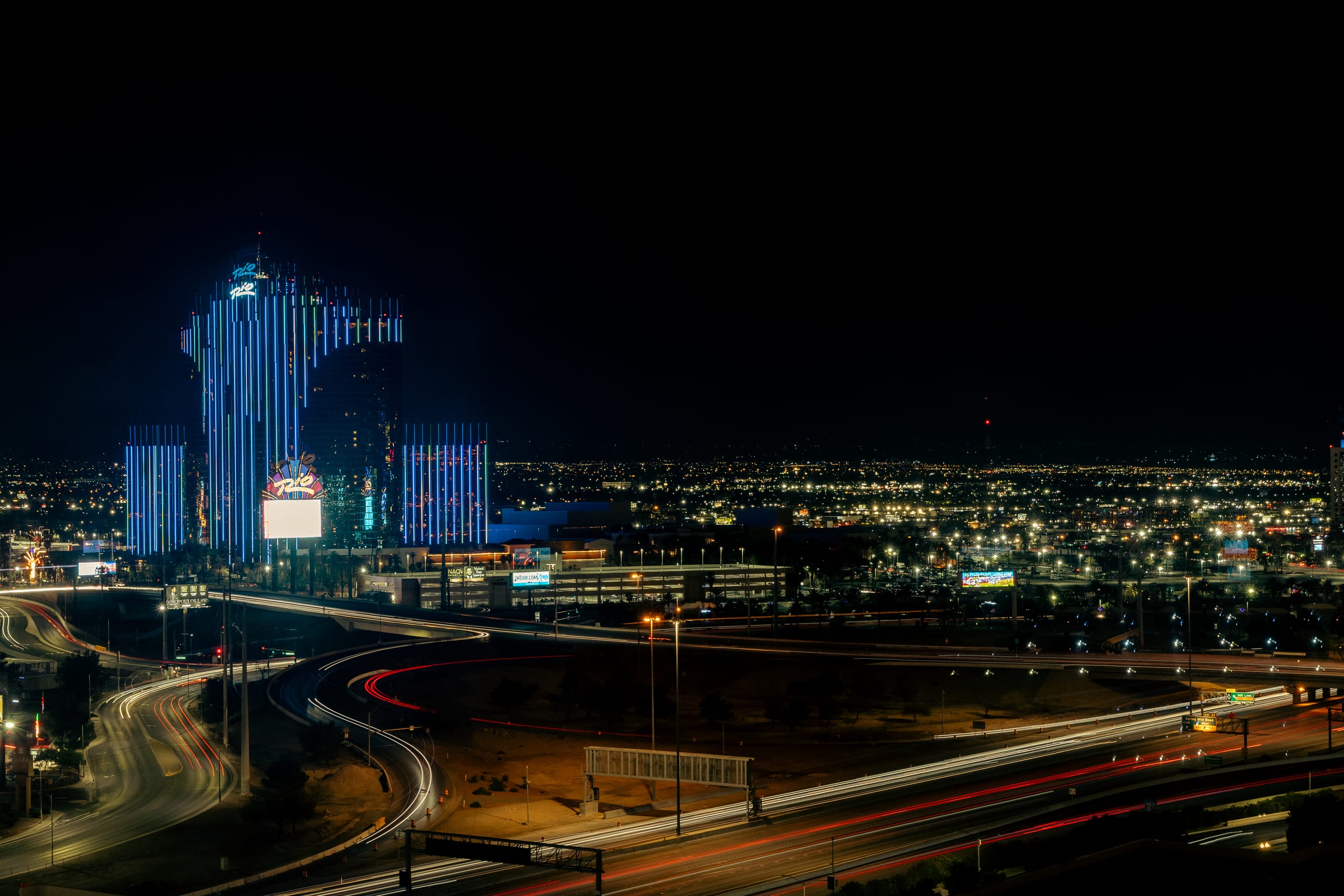 Rio hotel illuminated in blue above highway light trails at night